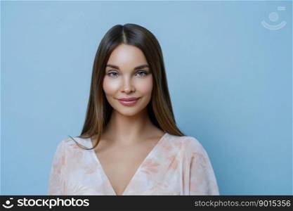 Beauty, youth, wellness and lifestyle concept. Pretty brunette Caucasian woman with beautiful hair and natural color, wears fashionable outfit, gazes at camera, isolated over blue background.