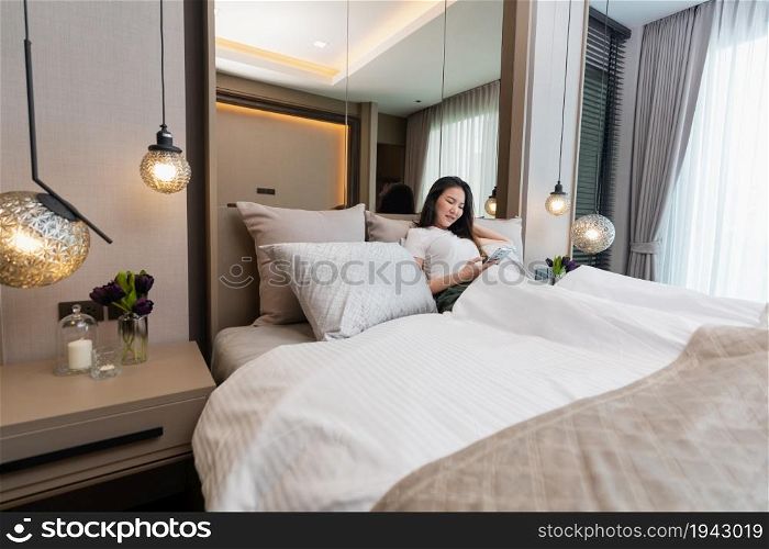 Bedroom concept a girl with drowsy feeling laying her back on several big and comfortable pillows on the bed looking on the mobile screen for social media.