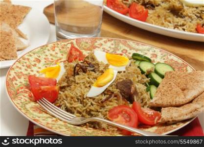Beef biryani served with tomato, cucumber, flat bread and a glass of water, with the serving dish int he background
