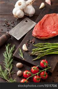 Beef braising steak, fresh raw slice on chopping board with garlic, asparagus and tomatoes with salt and pepper on wooden background with meat hatchet.
