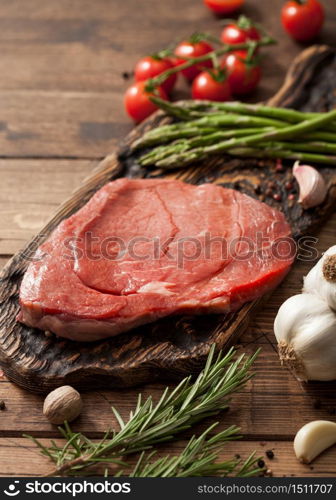 Beef braising steak, fresh raw slice on chopping board with garlic, asparagus and tomatoes with salt and pepper on wooden background. Macro