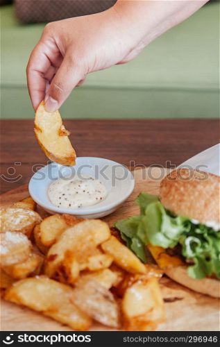 Beef cheeseburger and hand picking fried potato chunks with ranch dipping sauce on wood plate topside view shot