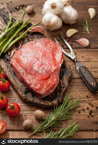 Beef fresh braising steak on chopping board with garlic, asparagus and tomatoes with salt and pepper with rosemary and meat fork on wooden background.
