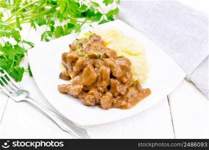 Beef goulash in tomato sauce with mashed potatoes in a plate, towel, parsley and fork on white wooden board background