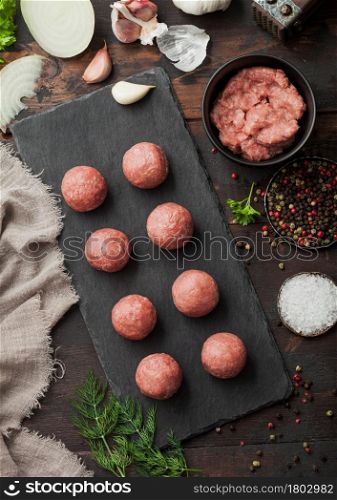 Beef meatballs on stone board with mince on bowl plate with pepper, salt and garlic on dark board with parsley and dill with onion.