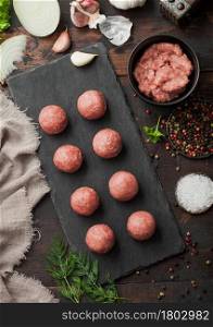 Beef meatballs on stone board with mince on bowl plate with pepper, salt and garlic on dark board with parsley and dill with onion.