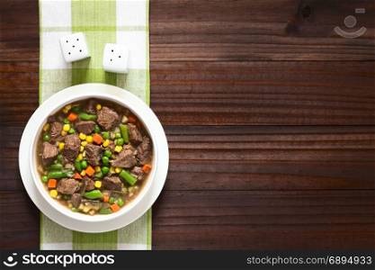 Beef stew or soup with colorful summer vegetables (pea, carrot, sweet corn, green bean, onion) in bowl, photographed overhead on dark wood with natural light. Beef Stew or Soup with Vegetables