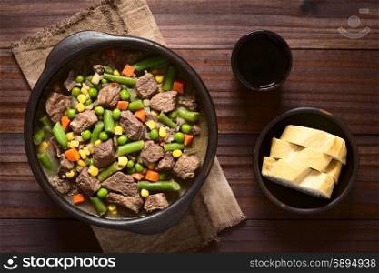 Beef stew or soup with colorful summer vegetables (pea, carrot, sweet corn, green bean, onion) in rustic bowl with red wine and bread slices on the side, photographed overhead on dark wood with natural light. Beef Stew or Soup with Vegetables