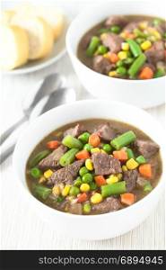 Beef stew or soup with colorful summer vegetables (pea, carrot, sweet corn, green bean, onion) in bowls with bread slices in the back, photographed with natural light (Selective Focus, Focus in the middle of the first dish). Beef Stew or Soup with Vegetables