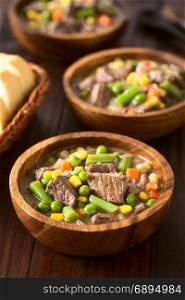 Beef stew or soup with colorful summer vegetables (pea, carrot, sweet corn, green bean, onion) in wooden bowls with bread slices on the side, photographed on dark wood with natural light (Selective Focus, Focus in the middle of the first dish). Beef Stew or Soup with Vegetables