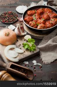 Beef traditional meatballs in tomato sauce in frying pan with pepper, garlic and parsley with onion and salt on wooden background.