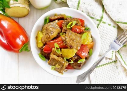 Beef with oranges, bell peppers and ginger root in bowl, a napkin and a fork on wooden board background from above