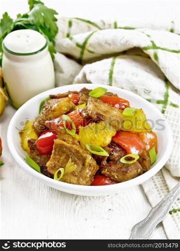 Beef with oranges, bell peppers and ginger root in bowl, a napkin and a fork on wooden board background