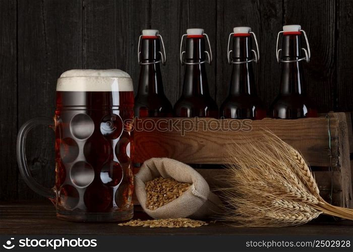 Beer glass on table with crate full of bottles, with brewing barley and wheat bunch, dark wooden background
