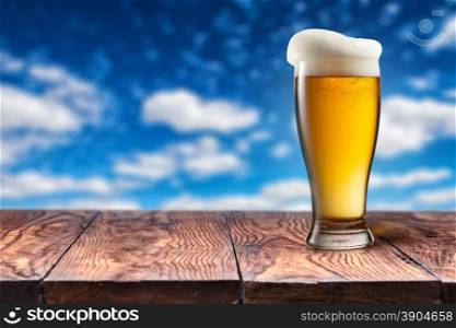 Beer in glass on wooden table against blue sky and clouds on natural background with bokeh. Beer in glass on wooden table against blue sky