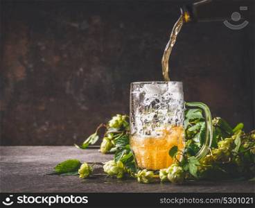 Beer is poured from a bottle into mug on dark rustic wooden background with hops, front view, copy space