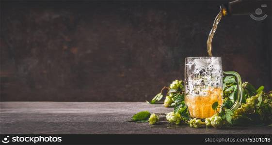 Beer is poured into mug on dark rustic wooden background with hops, front view, copy space, banner