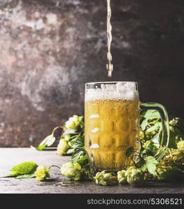 Beer pours into mug on table with fresh hops at dark rustic wall background, front view