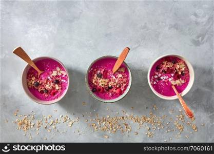 Beetroot smoothies with raspberries, granola, currants, almonds and sesame seeds in a plate with a wooden spoon on a gray concrete background with copy space. Healthy food. Top view. Delicious freshly prepared beetroot smoothies with raspberries, granola and sesame seeds in a plate with a spoon on a gray concrete background. Top view