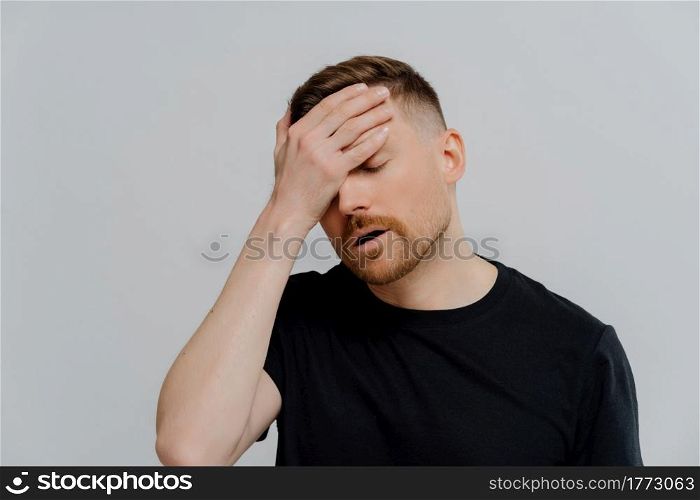 Being overworked. Young tired displeased ginger man in black shirt with closed eyes suffering from headache or mirgaine after hard working day, putting forehead and feeling sick. Worried man doing facepalm gesture and keeping eyes closed, concerning about troubles at work