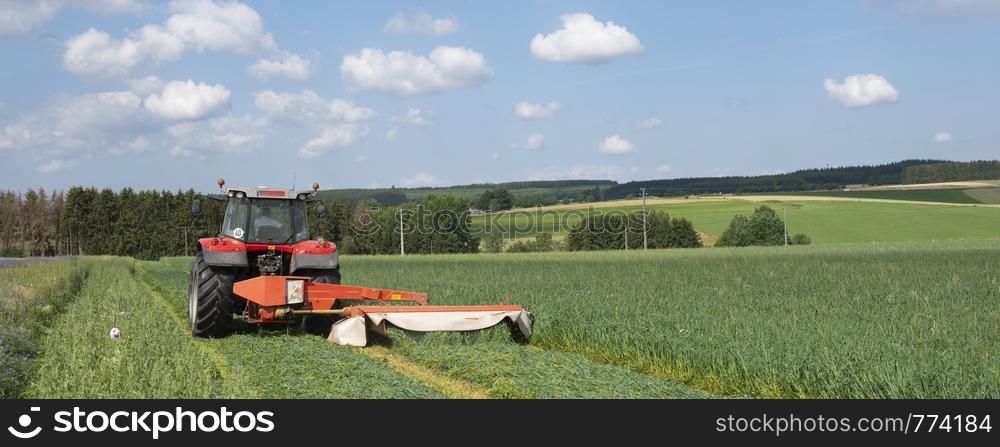 belgian farmer mows grass with tractor in field near Liege in the ardennes region on sunny summer day under blue sky
