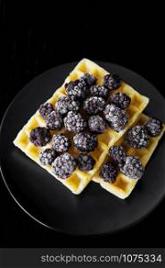 Belgian wafers with frozen blackberries on a black background, close-up.. Belgian waffles with blackberry