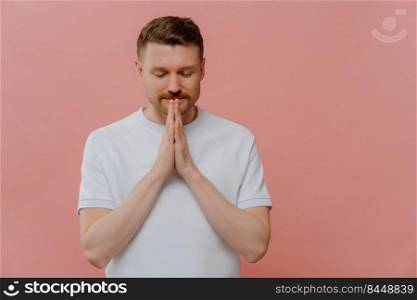 Believing in something good. Young handsome man in white t shirt keeping hands in praying gesture and eyes closed, asking for good luck or making wish while standing isolated over pink background. Young bearded man with closed eyes keeping hands in praying gesture