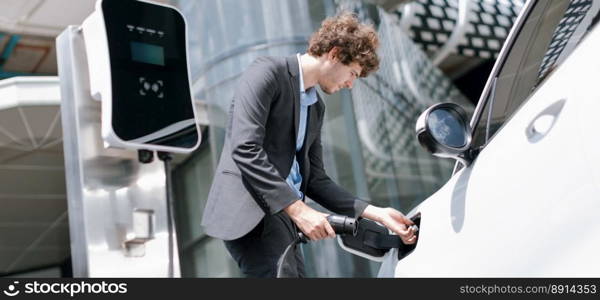 Below view closeup image of progressive black suit businessman recharge battery of his electric vehicle from public charging station. Renewable and alternative energy powered car concept.. Below view closeup progressive black suit businessman recharge battery of EV car