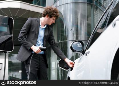 Below view closeup image of progressive black suit businessman recharge battery of his electric vehicle from public charging station. Renewable and alternative energy powered car concept.. Below view closeup progressive black suit businessman recharge battery of EV car