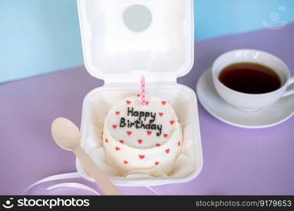 bento cake in eco-packaging with a wooden spoon and a candle next to a cup of tea on a purple and blue background. A small bento cake in eco-packaging with a wooden spoon and a candle next to a cup of tea on a purple and blue background