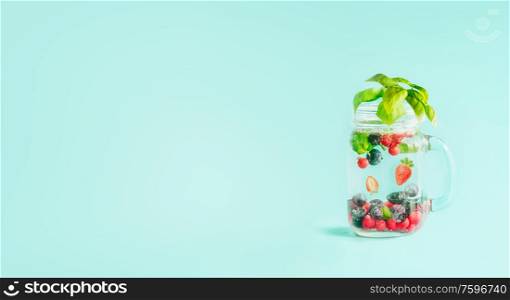 Berries detox fruit infused water in Mason jar flavored with herb leaves on table at sunny turquoise blue background. Summer mood drinks and lifestyle. Healthy background