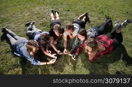 Best friends in circle social networking with mobile phones while lying on green grass in park. Top view. Wifi connected hipsters using smartphones. Many teenagers having fun in circle with cellphones surfing net and socializing. Technology addiction
