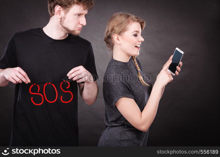 Betrayal and broken heart. Young blonde happy smiling woman flirting texting on mobile phone smartphone and sad depressed man with sos word sign symbol.. Blonde woman flirting texting on mobile phone.