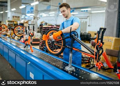 Bicycle factory, worker at assembly line, wheel installation. Male mechanic in uniform installs cycle parts in workshop, industrial manufacturing. Bicycle factory, assembly line, wheel installation