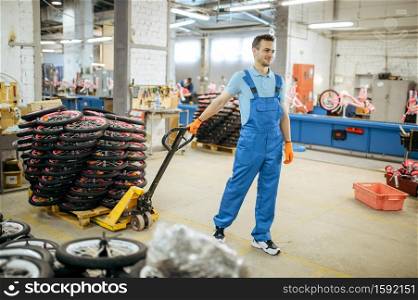 Bicycle factory, worker carries a cart with bike wheels. Male mechanic in uniform installs cycle parts, assembly line in workshop, industrial manufacturing. Bicycle factory, worker carries a cart with wheels