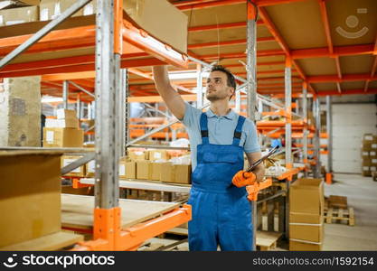 Bicycle factory, worker checks box on the shelf in bikes warehouse. Male mechanic in uniform at the packs with cycle parts, assembly line in workshop, industrial manufacturing. Bicycle factory, worker checks box on the shelf