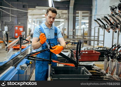 Bicycle factory, worker holds black teen bike frame. Male mechanic in uniform installs cycle parts, assembly line in workshop, industrial manufacturing. Bicycle factory, worker holds teen bike frame