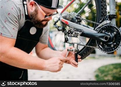 Bicycle mechanic hands adjusts bike chain with service tools. Cycle workshop outdoor. Bicycling sport, bearded repairman. Bicycle mechanic in apron adjusts bike chain