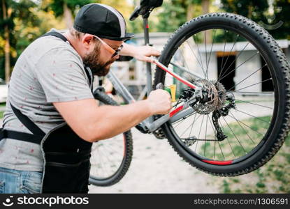 Bicycle mechanic in apron adjusts with service tools back disk brakes. Cycle workshop outdoor. Bicycling sport, bearded repairman