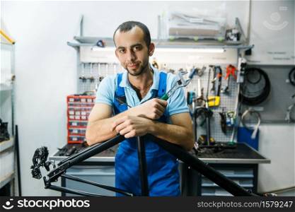 Bicycle repair in workshop, man poses at the frame. Mechanic in uniform fix problems with cycle, professional bike repairing service. Bicycle repair in workshop, man poses at the frame