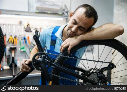 Bicycle repair in workshop, tired repairman sleeps at workplace. Mechanic in uniform fix problems with cycle, professional bike repairing service. Bike repair, tired repairman sleeps at workplace
