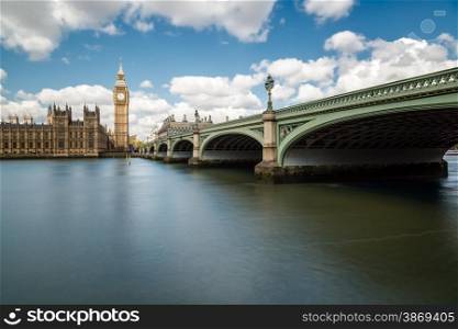 Big Ben and the Houses of Parliament in London with Westminster Bridge and the River Thames in the foreground against a blue sky with fluffy white clouds