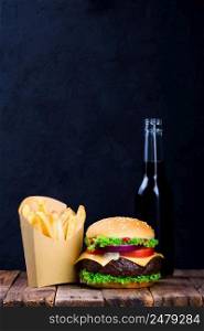 Big burger with french fries and drink on wooden table with dark stone background with copy space
