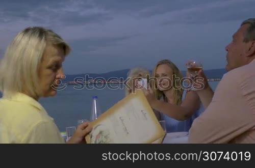 Big family enjoying evening in outdoor restaurant on the beach. Mother with adult daughter taking selfie, while other family members looking at menu and having wine