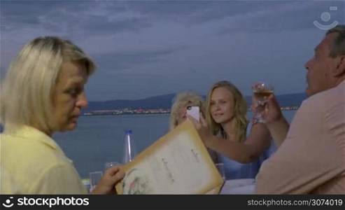 Big family enjoying evening in outdoor restaurant on the beach. Mother with adult daughter taking selfie, while other family members looking at menu and having wine