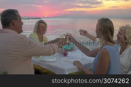 Big family in outdoor restaurant at the seaside at sunset. Adults clanging glasses with wine and child drinking water while waiting for food