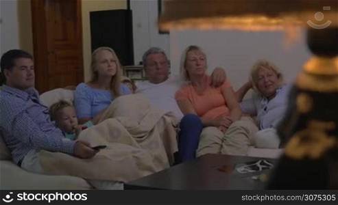 Big family spending evening at home. Parents, kid and grandparents sitting on big sofa and watching TV. Young woman feeling with events shown on the screen