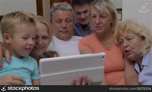 Big family with parents, son and grandparents watching video on digital tablet at home