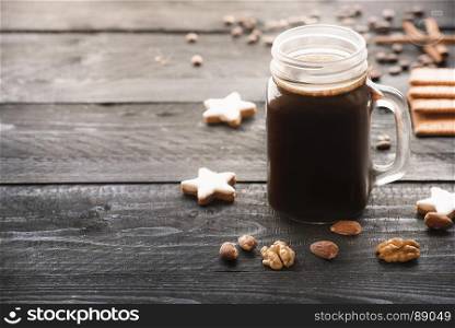 Big glass jar full with hot coffee and surrounded by cinnamon star shaped cookies, walnuts and almonds, on a rustic black wooden table, under the morning light.