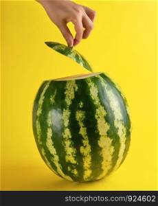 big ripe oval green watermelon and a female hand holds a cut piece, yellow background